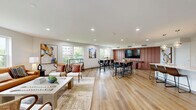 Located at 2305 Sokol Ct in Darien, Illinois, this single-floor community amenity space offers a well-designed, open-concept layout that seamlessly integrates a kitchen, dining area, and living room into one cohesive environment. Warm-toned hardwood-style flooring runs throughout, providing a consistent and inviting foundation for the entire space.

The kitchen features medium-toned wood cabinetry, a light quartz countertop peninsula with bar seating for three, stainless steel appliances, and globe pendant lighting overhead — creating a functional yet stylish hub for gathering. Adjacent to the kitchen, a high-top dining table with bar-height seating offers a casual space for meals or conversation, complemented by curated artwork and views to the outdoors.

The living area is bright and comfortable, furnished with a leather sofa, woven accent chairs, and a large coffee table, all bathed in natural light from multiple oversized windows that frame green outdoor views. A dedicated game zone includes a full shuffleboard table alongside a wall-mounted television, adding a recreational dimension to the space.

Floor-to-ceiling black-framed glass doors connect the lounge to an adjacent fitness center, offering visual openness and easy access. Recessed lighting throughout and neutral wall tones complete the clean, contemporary aesthetic of this thoughtfully appointed amenity space.