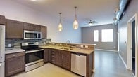 This single-floor space showcases a thoughtfully designed open-concept layout where the kitchen flows seamlessly into the living area. The kitchen is equipped with a full suite of stainless steel appliances, including a range, built-in microwave, and dishwasher. Dark espresso-toned cabinetry provides a rich contrast against the neutral-toned countertops, while a decorative mosaic tile backsplash adds visual texture and depth. An peninsula-style counter with an undermount double sink serves as a natural divider between the kitchen and living space, enhanced by two elegant amber glass pendant lights that hang overhead, casting warm ambient light. The living area features light hardwood-style flooring that extends throughout, contributing to a cohesive and airy feel. A ceiling fan is centrally positioned in the living space, adding both function and understated style. Two windows dressed with horizontal blinds allow natural light to filter into the space, complementing the cool gray wall tones. The overall ambiance is modern and clean, with a neutral palette and contemporary fixtures that create an inviting, functional environment.