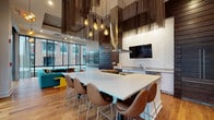 Located at 101–165 E Louisiana St in Indianapolis, this single-level communal space delivers a striking blend of modern design and urban sophistication. The layout is divided into two primary zones—a fully equipped kitchen and a comfortable lounge area—unified by warm hardwood flooring and a cohesive contemporary aesthetic throughout.

The kitchen area commands immediate attention with its dramatically tall ceilings adorned with cascading metallic chain curtains and a cluster of exposed Edison bulb pendant lights. A large, white-topped central island provides generous seating for multiple guests, while stainless steel appliances, a deep sink, and a wall-mounted flat-screen television complete the functional yet stylish setup. Dark slatted wood paneling and textured white brick-style wall treatments add visual depth to the space.

The adjoining lounge area is anchored by floor-to-ceiling windows that frame views of the surrounding Indianapolis urban streetscape, flooding the space with natural light. A bold teal sectional sofa, a wooden coffee table, and eclectic pendant lighting create an inviting, curated atmosphere. A raised platform with wooden steps and a black metal railing introduces architectural dimension, subtly delineating areas within the open plan. Together, these elements create a vibrant, well-appointed gathering space suited for socializing and entertaining.