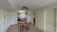 This single-floor residence offers a practical and functional layout centered around an open dining and kitchen area. The dining space comfortably accommodates a four-seat table and chairs, positioned beneath a ceiling fan with a light fixture that provides both air circulation and ambient lighting. The textured ceiling adds a classic residential character throughout.

The kitchen is efficiently arranged with wooden cabinetry, a white refrigerator, a range with an oven, and a sink positioned beneath a window that allows natural light to brighten the cooking area. The neutral-toned carpeted flooring flows seamlessly through the dining area, contributing to a warm and cohesive feel.

A hallway visible from the dining area leads to additional rooms, with white paneled doors providing access to closets and adjoining spaces. A glimpse into the connecting room reveals additional living or bedroom space, suggesting a well-connected floor plan. The overall color palette is light and neutral, with white walls and warm wood-toned cabinetry creating a clean and inviting atmosphere. The space is designed with practicality in mind, making efficient use of its footprint while maintaining a comfortable and livable environment throughout.