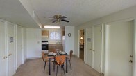 This single-floor residence offers a practical and functional layout centered around an open dining and kitchen area. The dining space comfortably accommodates a four-seat table and chairs, positioned beneath a ceiling fan with a light fixture that provides both air circulation and ambient lighting. The textured ceiling adds a classic residential character throughout.

The kitchen is efficiently arranged with wooden cabinetry, a white refrigerator, a range with an oven, and a sink positioned beneath a window that allows natural light to brighten the cooking area. The neutral-toned carpeted flooring flows seamlessly through the dining area, contributing to a warm and cohesive feel.

A hallway visible from the dining area leads to additional rooms, with white paneled doors providing access to closets and adjoining spaces. A glimpse into the connecting room reveals additional living or bedroom space, suggesting a well-connected floor plan. The overall color palette is light and neutral, with white walls and warm wood-toned cabinetry creating a clean and inviting atmosphere. The space is designed with practicality in mind, making efficient use of its footprint while maintaining a comfortable and livable environment throughout.