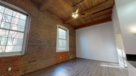Located at 2 N Main St in Beacon Falls, Connecticut, this thoughtfully converted single-floor space masterfully balances historic industrial character with modern updates. Original exposed brick walls run throughout, lending warmth and texture that speak to the building's storied past. Soaring ceilings with richly toned wood planking and heavy timber beams create a dramatic overhead canopy, while ceiling fans enhance comfort in the open living areas.

The main living area features durable hardwood-style flooring that reflects the ample natural light streaming through large, multi-pane windows with classic arched detailing. A secondary carpeted room, also anchored by exposed brick and generous windows, offers a quieter, more intimate atmosphere with views of the surrounding streetscape.

The kitchen is well-appointed with warm maple cabinetry, dark granite countertops, stainless steel appliances including a refrigerator, range, and microwave, and tile flooring. A peninsula counter opens the kitchen toward the living space, reinforcing the open-concept flow. The soaring ceiling volume in the main living area provides an airy, loft-like quality throughout.

Together, the preserved architectural details and updated interior finishes create a distinctive and functional space that honors its historic roots while offering modern comfort in the heart of Beacon Falls.