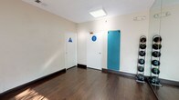 This single-floor fitness studio is thoughtfully designed to support a variety of workout activities in a clean, functional environment. The main exercise area features rich, dark hardwood flooring that runs throughout, providing a durable and attractive surface well-suited for physical activity. Large floor-to-ceiling mirrors line one wall, visually expanding the space and allowing users to monitor their form during workouts. Natural light streams in through multiple windows fitted with vertical blinds, as well as a glass-paneled exterior door that opens directly to an outdoor area, offering both ventilation and a bright, welcoming atmosphere. Recessed and flush-mounted ceiling light fixtures provide consistent overhead illumination. The studio is equipped with wall-mounted exercise mats in a distinctive teal color and freestanding medicine ball racks stocked with a full range of weighted balls, supporting strength and functional training. Separate restroom facilities — marked with clearly visible gender-specific signage — are conveniently accessible from within the main workout space. The neutral, cream-toned walls keep the aesthetic clean and distraction-free, while the overall layout remains open and flexible to accommodate individual training sessions or small group fitness classes.
