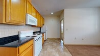 This single-floor unit is thoughtfully laid out to maximize functionality and comfort. The kitchen is a standout feature, equipped with warm honey-toned wood cabinetry extending to the ceiling, dark countertops, a subway tile backsplash, and a full suite of white appliances including a range, over-the-range microwave, dishwasher, and refrigerator. Light ceramic tile flooring anchors the kitchen and transitions seamlessly into the adjacent living areas.

The living space is open and bright, featuring plush wall-to-wall carpeting, neutral cream-toned walls, and a ceiling fan with integrated lighting. Large windows allow natural light to fill the room, while individual wall-mounted HVAC units provide climate control. The layout connects the living area to multiple carpeted rooms, each with their own windows fitted with horizontal blinds and individual HVAC units, providing both privacy and comfort.

The bathroom is clean and well-finished, featuring the same warm wood cabinetry with a dark countertop and undermount sink, a mirrored medicine cabinet with overhead vanity lighting, and tile flooring that complements the rest of the unit. Overall, the space presents a cohesive, functional design with consistent finishes throughout.
