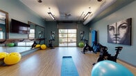 Located at 10315 Monrovia Drive in Matthews, NC, this single-floor fitness and yoga studio is thoughtfully designed to support a wide range of exercise and wellness activities. The space features light hardwood-style flooring throughout, which provides a clean and practical surface ideal for yoga, stretching, and group fitness classes.

The studio is equipped with multiple spin bikes arranged for group cycling sessions, a variety of stability and exercise balls in assorted sizes and colors, yoga mats, and a wall-mounted medicine ball rack. A full-length large mirror with a warm wood frame spans one wall, accompanied by a barre rail — ideal for stretching and balance exercises.

The walls are painted in a calming teal-blue hue, complemented by bold, artistic portrait paintings that lend the space a creative and energizing atmosphere. A decorative textured accent wall near the entrance adds visual interest and depth to the overall design.

Natural light fills the studio through a large glass garage-style door, which opens to an outdoor view and creates a bright, airy ambiance. Overhead, industrial-style pendant lighting and a ceiling fan ensure comfort and visibility during workouts. Exposed ductwork contributes to the studio's modern, industrial aesthetic. Studio rules are posted on the wall, and the space is equipped with climate control for year-round comfort.