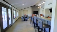 This single-floor space features a contemporary open-plan layout designed for comfortable gatherings and socializing. The interior is finished with light hardwood-style flooring that extends throughout, complementing the neutral-toned walls accented by a darker gray feature wall. Vaulted ceilings with exposed beam detailing add architectural interest and contribute to an airy, open feel, while recessed lighting ensures even illumination across the space.

A prominent bar counter runs along one side of the room, lined with tall, gray upholstered bar stools featuring nailhead trim detailing, providing ample seating for guests. An elegant chandelier with cylindrical pendant shades hangs above the bar area, adding a refined decorative touch. A large mirror positioned behind the bar enhances the sense of depth and light throughout the space.

In the main gathering area, round black pedestal tables are surrounded by modern gray accent chairs, creating an inviting setting for casual dining or conversation. A wall-mounted flat-screen television is positioned on the feature wall, making the space well-suited for entertainment. Natural light enters through multiple windows along the exterior wall, further brightening the overall ambiance.