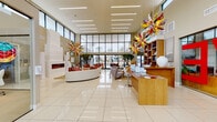 This single-floor amenity space makes a bold first impression with a grand, light-filled lobby distinguished by soaring ceilings and expansive floor-to-ceiling glass walls that flood the interior with natural light and offer open views of the outdoor surroundings. Gleaming large-format tile flooring extends throughout, reflecting the bright, airy atmosphere. Dramatic multi-colored starburst chandelier sculptures serve as eye-catching artistic focal points suspended from the ceiling. The lobby features a curved lounge seating arrangement centered around a sleek linear fireplace set into a textured stone wall, creating an inviting social hub. A warm wood reception desk anchors the space alongside built-in shelving displays. Indoor trees add organic texture, while sliding glass doors connect seamlessly to an outdoor courtyard furnished with colorful seating. The fitness center is comprehensively equipped with cardio machines, cable systems, free weights, medicine balls, kettlebells, and cycling bikes, all set against bold red accent walls and floor-to-ceiling mirrors that amplify the space. An adjacent studio area with hardwood flooring provides a dedicated zone for group fitness. A separate wellness or salon room features white sculpted tile walls, warm wood cabinetry, and curated contemporary artwork, offering a refined and tranquil environment.