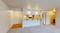 This single-floor residence offers a practical and comfortable layout designed for everyday living. The open-concept living area is finished with plush wall-to-wall carpeting and connects seamlessly to the kitchen via a peninsula-style breakfast bar with granite-look countertops and built-in shelving brackets — ideal for casual dining or entertaining. The kitchen is fully equipped with warm honey-toned cabinetry, a suite of white appliances including a refrigerator, microwave, and electric range, and is illuminated by recessed accent lighting. Hardwood-style flooring runs through the entry hallway and kitchen, providing a clean contrast to the carpeted living spaces. A dedicated in-unit laundry area is conveniently located just off the kitchen, housing a full-sized washer and dryer. The bedrooms are generously sized with soft carpeting, neutral warm-toned walls, and windows fitted with horizontal blinds that allow for natural light and outdoor views of surrounding greenery. One bedroom features a sliding closet door. The bathroom includes a wide vanity with warm wood cabinetry, a large mirror, light bar, and a full tub-shower combination. The living room opens to an outdoor area through a sliding glass door framed by vertical blinds, adding natural light and a sense of openness to the space.