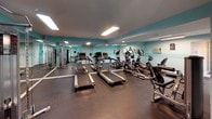 This fitness center is designed to provide a comprehensive workout environment within a single-level layout. The space is styled with a distinctive two-tone teal and white color scheme along the walls, paired with a dark ceiling that lends a modern, energetic atmosphere to the room. Durable rubber flooring covers the entire workout area, providing both safety and stability during exercise.

The cardio section is well-stocked, featuring multiple treadmills and elliptical trainers arranged in rows, offering ample options for aerobic training. A cable machine provides strength-training functionality, rounding out the equipment offerings for a balanced fitness experience. Full-length mirrors line one wall, visually expanding the space while allowing users to monitor their form during workouts. Recessed and flush-mounted fluorescent lighting ensures the room is well-illuminated throughout.

A wall-mounted air conditioning unit maintains a comfortable temperature within the workout area. An adjacent entryway, finished in neutral tones with tile flooring and a branded welcome mat, leads to glass-paneled doors that open to an outdoor area, allowing natural light to filter into the transition space. Posted fitness center rules are clearly displayed near the entrance.