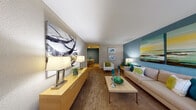 This single-floor residence is thoughtfully designed with a cohesive color palette of teal, green, and neutral tones that carries throughout every room. The living area serves as the focal point of the home, featuring a bold teal accent wall adorned with large-scale artwork, plush carpeting, and ample natural light streaming through a wide window and entry door. A long, low credenza with table lamps anchors one side of the room, complementing the comfortable seating arrangement centered around a minimalist coffee table. The living area flows openly into the dining space, which is highlighted by a warm yellow accent wall and a casual dining set, creating an inviting atmosphere for entertaining. The galley-style kitchen is efficiently laid out with white cabinetry, countertop workspace on both sides, tile flooring, and full appliances including a range and refrigerator, all set against a soft sage green accent wall. The residence includes two carpeted bedrooms, each equipped with a ceiling fan and window treatments that allow for natural light. The primary bedroom features its own teal accent wall, while the secondary bedroom offers a dedicated desk area. A full bathroom with a vanity, white cabinetry, and tile flooring provides practical functionality. A carpeted hallway connects the main living spaces to the private areas of the home.
