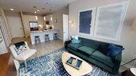 This single-floor space showcases an open-concept layout that seamlessly connects the living area and kitchen, creating a cohesive and functional environment. The living area is anchored by a tufted emerald green sofa paired with teal and coral accent pillows, positioned atop a bold blue geometric area rug. A rounded wood coffee table serves as a central focal point, while a wall-mounted sconce and two large abstract navy blue art pieces add visual interest to the neutral-toned walls.

The kitchen flows directly from the living space and features a peninsula-style breakfast bar with sleek bar stools, providing both casual dining and additional workspace. White cabinetry with light countertops creates a clean, bright aesthetic, while pendant lighting adds a modern touch above the bar. Stainless steel appliances, including a full-sized refrigerator and microwave, are integrated seamlessly into the cabinetry. Light wood-style flooring runs throughout, tying the two areas together and enhancing the sense of openness. Recessed ceiling lighting provides ample illumination across the entire space.