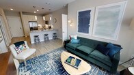 This single-floor space showcases an open-concept layout that seamlessly connects the living area and kitchen, creating a cohesive and functional environment. The living area is anchored by a tufted emerald green sofa paired with teal and coral accent pillows, positioned atop a bold blue geometric area rug. A rounded wood coffee table serves as a central focal point, while a wall-mounted sconce and two large abstract navy blue art pieces add visual interest to the neutral-toned walls.

The kitchen flows directly from the living space and features a peninsula-style breakfast bar with sleek bar stools, providing both casual dining and additional workspace. White cabinetry with light countertops creates a clean, bright aesthetic, while pendant lighting adds a modern touch above the bar. Stainless steel appliances, including a full-sized refrigerator and microwave, are integrated seamlessly into the cabinetry. Light wood-style flooring runs throughout, tying the two areas together and enhancing the sense of openness. Recessed ceiling lighting provides ample illumination across the entire space.