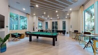 Located at 2810 University Square Drive in Tampa, Florida, this single-floor amenity lounge offers a well-appointed recreational and social space designed with a contemporary aesthetic. The game room serves as the centerpiece, featuring a full-size pool table with green felt, a regulation shuffleboard table set against a rich dark wood accent wall, and a foosball table — all set atop light-toned herringbone-patterned flooring that runs throughout the space. Decorative pendant lighting, including a distinctive geometric multi-bulb fixture, adds a stylish ambiance to the game area. Large floor-to-ceiling windows and glass doors flood the interior with natural light and provide open views of the surrounding outdoor courtyard with tropical landscaping. A casual dining area is equipped with modern high-top tables and sleek metal-framed chairs, complemented by pendant lighting overhead. Additional lounge seating with bistro-style high-top tables is positioned near the expansive windows. A wall-mounted flat-screen television offers entertainment options alongside the game tables. The overall design balances a clean, modern interior palette of neutral tones with bold accent furnishings, creating an inviting and versatile community gathering space.