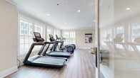 Located at 180 Westfall Town Dr in Matamoras, PA, this single-floor fitness center is thoughtfully designed to support a comprehensive workout experience. The space is divided into distinct functional zones, each outfitted with professional-grade equipment and finished with light wood-look flooring and neutral warm-toned walls that create a clean, welcoming atmosphere.

The cardio area is generously proportioned, featuring multiple treadmills, elliptical trainers, and a stair climber arranged along a bank of large, multi-pane windows. These windows flood the room with natural light and offer views of the surrounding community, including a glimpse of the outdoor pool area, making cardio sessions feel open and energizing.

A separate strength and functional training room houses a full power rack with bench press, a cable machine, a spin bike, and a well-stocked equipment rack loaded with resistance bands, kettlebells, medicine balls, and additional accessories. Recessed lighting, crown molding, and a wall-mounted climate control unit add a polished, comfortable feel to this area.

Throughout both zones, the space maintains a bright, airy ambiance supported by recessed ceiling lights. The layout allows for ample floor space between equipment, ensuring ease of movement and a safe training environment. The overall design balances practicality with a refined aesthetic befitting a modern community fitness facility.