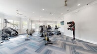 Located at 920 Yorkmont Ridge Lane in Fort Mill, South Carolina, this single-floor fitness center offers a well-appointed and thoughtfully designed space for residents to pursue their wellness goals. The main workout area is generously proportioned, featuring a striking herringbone-patterned carpet in a blue and grey palette that adds a modern, polished aesthetic throughout. Recessed lighting and ceiling fans maintain a bright and comfortable environment, while elegant crown molding lends a refined architectural touch. Large windows along multiple walls flood the space with abundant natural light and offer views of the surrounding greenery, creating an inviting atmosphere. The facility is equipped with a comprehensive selection of cardio machines, including treadmills, ellipticals, stair climbers, stationary bikes, rowing machines, and a weight rack with plates, catering to a full range of fitness routines. Wall-mounted flat-screen televisions provide entertainment during workouts. A separate, carpeted flex room adjacent to the main floor features its own windows, wall-mounted mirror or display panel, and chair rail detailing, making it suitable for stretching, yoga, or other activities. The overall design balances functionality with an upscale aesthetic, offering a clean, bright, and welcoming fitness environment.