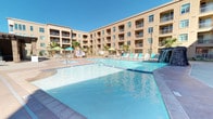 This property showcases an impressive outdoor amenity complex designed for relaxation and recreation. At its centerpiece are two distinct pool areas — one nestled within a courtyard framed by a multi-story stucco building with private balconies, and another positioned against a dramatic red rock desert landscape. Both pools are generously sized, featuring crystal-clear blue water, dedicated hot tubs, and ample deck space lined with turquoise lounge chairs. A decorative waterfall feature adds a resort-like ambiance to the courtyard pool area, which is further enhanced by swaying palm trees and shaded seating under green umbrellas. The second pool area benefits from a covered pergola structure and an open-air cabana with additional seating, offering sweeping views of the surrounding desert terrain and red rock formations. A covered lounge area provides a comfortable indoor-outdoor retreat, furnished with wicker seating accented by vibrant teal cushions and equipped with vending machines for added convenience. Soaring ceilings and dappled natural light filtering through the overhead structure give this lounge a breezy, welcoming character.