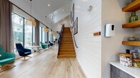 Located at 347 Arboretum Dr in Wilmington, NC, this community clubhouse is a thoughtfully designed gathering space that blends modern coastal aesthetics with functional amenities. The architecture is defined by dramatic vaulted ceilings with exposed wood beam accents, floor-to-ceiling black-framed windows, and light hardwood flooring that runs throughout the space, creating a bright, airy atmosphere.

The main lounge area features an expansive seating arrangement with contemporary accent chairs in rich jewel tones — teal, green, and charcoal — paired with round pedestal tables and woven pendant lighting. A wall of oversized windows floods the space with natural light and offers views of the surrounding landscaping and community exterior.

A second seating zone provides a relaxed living room feel with a plush sofa, woven chairs, a sculptural white coffee table, and a patterned area rug, all set against full-length glass doors that open to outdoor views. A dedicated bar-height window ledge with woven rattan stools offers an additional perch overlooking the pool area.

The kitchenette is outfitted with sleek wood-grain cabinetry, white quartz countertops, a matte black undermount sink, a gold pull-down faucet, and a striking geometric tile backsplash. A staircase with warm wood treads and a black metal railing leads to an upper mezzanine level, anchoring the double-height entry with visual drama. White shiplap wall cladding ties the design together with a clean, coastal-modern finish.