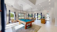 Located at 2450 Lake Debra Dr in Orlando, FL, this single-floor community clubhouse is a well-appointed gathering space designed for recreation and socializing. The room features soaring ceilings with recessed lighting and a coffered ceiling detail that adds architectural interest overhead. Large light-colored porcelain tile floors run throughout, enhancing the bright, airy atmosphere.

A full-size pool table anchors the central area, positioned atop a decorative area rug, making it an immediate focal point. Adjacent to the game area, a comfortable lounge zone features wood-framed seating with white cushions and lime green accent pillows, arranged around a round wood coffee table. The lounge benefits from abundant natural light streaming through oversized windows and a glass panel garage-style door that opens to the outdoor courtyard, offering views of the lushly landscaped grounds and the community's Mediterranean-style architecture.

A bar-height counter with seating runs along one wall, ideal for casual gatherings. A wall-mounted television and a decorative clock gallery add personality to the space. A glass-enclosed fitness center is visible from the main area, providing a glimpse of exercise equipment within. Navy blue drape accents throughout tie the design together, creating a cohesive, resort-inspired ambiance.