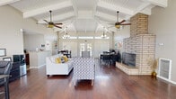 Located in Palmdale, CA, this single-floor space makes a striking first impression with its dramatic vaulted ceilings adorned with exposed white-painted wood beams and planking, lending an airy, open character throughout. Multiple ceiling fans and elegant chandelier-style pendant lights are suspended from the beamed ceiling, ensuring both comfort and ambiance. Rich, dark hardwood flooring runs throughout, providing a warm contrast to the bright, neutral walls and ceiling.

The main open area serves as both a lounge and common gathering space, furnished with a sectional sofa, patterned accent chairs, and a round side table arranged around a bold, floor-to-ceiling brick fireplace that anchors the room as its central architectural feature. A workstation with desks and office equipment is integrated into the open floor plan, offering a functional workspace within the communal setting.

The dining area is defined by a dark wood dining table and chairs set beneath a modern multi-bulb chandelier, bathed in natural light from large windows with slatted blinds. French doors along the main wall flood the space with additional daylight and provide views to the outdoors.

A dedicated exercise room, accessible directly from the main area, is equipped with fitness machines, catering to an active lifestyle. The overall ambiance is bright, welcoming, and versatile, blending comfort with functionality.