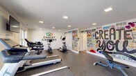Located at 320 W Branch Ave in Pine Hill, NJ, this single-floor fitness facility offers a well-organized and functional layout designed to support a variety of workout routines. The space is divided into three distinct areas: a main exercise room, a yoga and stretching room, and a full bathroom.

The main exercise room is the heart of the facility, featuring rubber flooring ideal for high-impact activity. It is equipped with multiple treadmills, an elliptical machine, a stationary bike, and a multi-use cable weight machine. A wall-mounted flat-screen TV is positioned between two large windows that bring in natural light and views of the surrounding greenery. A bold, colorful graffiti-style mural declaring "CREATE" energizes the space, while a mirrored wall enhances the sense of openness. Recessed lighting provides consistent, even illumination throughout.

Adjacent to the main gym is a dedicated yoga and stretching room with warm wood-look flooring, clean white walls, and minimalist botanical artwork. Yoga mats are laid out and ready for use, and the calm, bright atmosphere makes it well-suited for low-impact exercise and mindfulness practices.

The bathroom is clean and practical, with wood-look flooring, white walls, a pedestal sink, and a full bathtub with a shower curtain — providing convenient post-workout amenities.