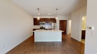 This single-floor residence offers a thoughtfully designed open-concept layout that seamlessly connects the kitchen and living areas. The kitchen is a standout feature, anchored by a large central island with a white countertop and an undermount sink, complemented by elegant pendant lighting above. Rich dark-espresso cabinetry lines the walls, providing ample storage, while a classic white subway tile backsplash adds a crisp contrast. Stainless steel appliances, including a refrigerator, range, and built-in microwave, complete the well-equipped culinary space. Warm wood-tone flooring runs throughout the open living area, lending a cohesive and inviting atmosphere. The bedroom is carpeted for comfort and features a ceiling fan and a large window that fills the room with natural light and outdoor views, creating a bright and airy retreat. The bathroom is finished with neutral large-format tile flooring, a dark-cabinet vanity with a white countertop, and accessible grab bars, reflecting both style and practicality. Clean white walls and neutral tones throughout the residence create a calm, modern ambiance that is both functional and visually cohesive.