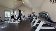 This fitness center is thoughtfully designed to support a comprehensive workout experience within a bright, open single-level layout. The space is defined by its striking vaulted ceilings, which lend an airy, spacious feel throughout. Large multi-pane windows line multiple walls, flooding the interior with natural light and offering views of surrounding greenery and outdoor areas.

The flooring throughout is durable dark rubber, providing traction and cushioning suited for high-activity use. The cardio zone is well-appointed with multiple treadmills and a stationary bike, positioned to take advantage of the natural light and wall-mounted television. A motivational fitness-themed word art mural adds visual character to one accent wall.

The strength training area includes a multi-station cable machine, a weight bench, and a full rack of dumbbells in graduated sizes. A water cooler station provides convenient hydration access, and a wall clock keeps users on schedule.

Recessed lighting and a ceiling fan supplement the natural light, ensuring comfortable conditions at all hours. The neutral gray walls and clean white trim create a modern, uncluttered aesthetic that keeps the focus on functionality and performance.