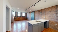 Located at 356 Hertel Ave in Buffalo, NY, this two-floor loft seamlessly blends industrial character with contemporary updates. The main level hosts an open-concept combined living and dining area that flows directly into the kitchen, creating a spacious and connected living environment. The kitchen features a central island with a white quartz countertop, undermount sink, and sleek gray cabinetry, accented by modern pendant lighting and a black track lighting system overhead. Warm hardwood flooring runs throughout both levels, complementing the richly textured exposed brick walls that define the loft's industrial aesthetic. A standout patina metal barn door adds an artful architectural focal point to the main living area, which is also filled with natural light from large multi-pane windows. A modern open staircase with black steel railings leads to the upper level, where the generously sized primary bedroom resides alongside two closets. The upper floor retains the industrial character with exposed wooden beam ceilings, visible ductwork, and continued brick wall detailing. The full bathroom features large-format white tile flooring, a floating vanity, and a continued exposed brick accent wall for design cohesion. A dedicated laundry area adds everyday convenience to this thoughtfully designed urban loft.