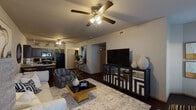 This two-bedroom, two-bathroom single-story unit features an open-concept design that seamlessly connects the living room, dining area, and kitchen. The living room is generously proportioned, showcasing dark hardwood-style flooring, a ceiling fan with light fixture, and large windows fitted with horizontal blinds that allow natural light to filter in. A stylish media console anchors one wall, while the layout provides ample space for comfortable seating arrangements.

The kitchen is equipped with dark wood cabinetry, light-toned countertops, and black appliances including a range, built-in microwave, dishwasher, and refrigerator. A breakfast bar with barstool seating provides a casual dining option directly adjacent to the kitchen.

Both bedrooms are well-sized, featuring plush carpeting, ceiling fans, and windows that bring in natural light. Each bedroom is served by its own full bathroom. The bathrooms include dark wood vanity cabinetry, framed mirrors, and one features a tub/shower combination with sliding glass doors. Wall-mounted storage cabinets in both bathrooms offer additional organizational space. The overall ambiance of the unit is warm and contemporary throughout.