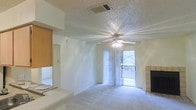 This single-floor residence centers around an open living area that is both functional and inviting. The room is finished with wall-to-wall carpeting in a neutral tone, contributing to a soft, comfortable atmosphere throughout the space. A textured popcorn ceiling adds dimension overhead, while a ceiling fan with an integrated light fixture provides both ventilation and ambient lighting.

A tiled fireplace anchors one side of the living area, serving as a notable architectural feature that adds warmth and character to the room. On the opposite end, sliding glass doors framed by vertical blinds open directly onto a private balcony, allowing natural light to flood the interior and offering pleasant views of surrounding greenery.

An adjacent room, partially visible through an open doorway, suggests a connected bedroom or secondary living space, reinforcing the open and accessible layout of the floor plan. The overall design emphasizes simplicity and practicality, with neutral walls and understated finishes that provide a versatile backdrop. The combination of indoor comfort and outdoor access makes this space well-suited for everyday living.