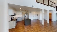 This single-floor space showcases a thoughtfully designed open-concept layout with notable architectural height, as seen in the dramatic vaulted ceiling above the main living and kitchen area. Rich hardwood flooring runs consistently throughout, lending warmth and continuity to each room.

The kitchen serves as a focal point of the space, centered around a sizeable island with dark granite countertops and a built-in sink. Stainless steel appliances, including a French door refrigerator, electric range, and microwave, are complemented by two-tone cabinetry featuring white upper cabinets and gray lower cabinets. White subway tile backsplash and pendant lighting complete the contemporary aesthetic.

The open floor plan allows the kitchen to flow seamlessly into the living area, supported by structural columns that add architectural character. A mezzanine-style overlook is visible above, adding further visual depth to the space.

The bathroom features a modern floating vanity with granite countertops, backlit mirror, and clean white finishes. A dedicated laundry room provides side-by-side washer and dryer units, an overhead wooden rod for hanging, and ample floor space, all finished with matching hardwood flooring.