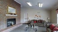 This single-floor space centers around a welcoming living area that combines classic and contemporary design elements. A prominent full-height brick fireplace wall serves as a striking focal point, complete with a traditional wood mantle and raised brick hearth, adding warmth and architectural character to the room. The fireplace surround's rich, varied brick tones provide a bold contrast against the smooth, neutral-toned walls.

The living area is carpeted in a patterned grey-blue tone that extends throughout the space, adding texture underfoot. A large, cushioned sofa dressed with red and patterned accent pillows anchors the seating area, positioned around a glass-topped coffee table with an ornate metal base. Decorative sculptural pieces and vases rest on the table, contributing to the room's styled aesthetic.

One wall features hand-painted floral wall art in warm oranges and greens, introducing a creative, personalized touch. Overhead, flush-mount fluorescent light fixtures provide consistent, even illumination throughout the space. A side table with a traditional lamp and a six-panel interior door are also visible, rounding out the functional and decorative elements of this inviting, well-appointed living space.