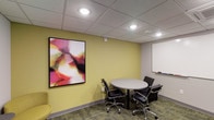 This single-floor meeting room is designed to facilitate focused, small-group collaboration in a compact yet functional setting. The space is defined by warm yellow-green walls that create an energizing atmosphere, complemented by a drop ceiling with a recessed grid panel system and evenly spaced recessed lighting that ensures consistent, well-distributed illumination throughout the room. At the center of the space sits a round gray table accompanied by black ergonomic rolling chairs, providing a comfortable and practical configuration ideal for intimate discussions or brainstorming sessions. A large whiteboard mounted on one wall serves as a key functional feature, offering a dedicated surface for note-taking and visual presentations. Adding a creative dimension to the room, a bold large-scale abstract artwork in vivid pinks, reds, and whites is prominently displayed on the adjacent wall, introducing an artistic focal point that enlivens the otherwise utilitarian environment. The flooring consists of patterned commercial carpet tiles in muted tones, contributing to the professional character of the space. Overall, the room balances practicality with a touch of visual interest, making it well-suited for professional meetings or collaborative work sessions.