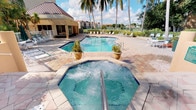 This outdoor amenity space embodies a classic Florida resort aesthetic, centered around a generously sized rectangular swimming pool with clear, blue-tinted water. Adjacent to the pool sits a large octagonal spa, complete with active jets that keep the water in constant, relaxing motion — accessible via stainless steel handrails. The surrounding pool deck is finished in warm-toned, patterned tiles that complement the tropical atmosphere, and is outfitted with multiple lounge chairs positioned to take advantage of the abundant natural sunlight.

Tall, mature palm trees frame the space, providing both visual character and partial shade while reinforcing the lush, tropical ambiance. Potted ornamental grasses add greenery at ground level near the pool's edge. A covered structure adjoins one side of the pool area, offering a shaded retreat from the sun. The surrounding landscape is dense with tropical foliage, including manicured shrubs and additional palms that create a sense of privacy and seclusion.

Views from the space extend toward lush greenery and open sky, enhancing the open-air, resort-like feel. The overall setting is bright, airy, and well-maintained, making it a welcoming outdoor destination suited for both relaxation and recreation.