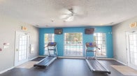 This fitness center is a comprehensively equipped exercise facility designed to support a full range of workout routines. The space is laid out across a single floor and is divided into functional zones, each catering to different types of training.

The cardio area is a standout feature, characterized by its vibrant teal accent wall and large windows that overlook an outdoor pool area, allowing natural light to fill the space and providing an engaging view during workouts. Multiple treadmills are positioned facing the windows, and wall-mounted flat-screen televisions are installed above for entertainment. A ceiling fan overhead aids in air circulation during exercise sessions.

Adjacent areas house additional cardio equipment, including an elliptical trainer and stationary spin bikes, arranged on durable rubber flooring that extends throughout the facility. The walls in these sections are finished in a neutral gray tone, contributing to a clean, focused atmosphere.

A dedicated strength training zone features a multi-function cable machine and ample open floor space. Full-length mirrors line one wall, enabling users to monitor their form. The textured ceiling and recessed lighting provide consistent illumination throughout. An exit door with glass panels connects the space to the exterior, ensuring convenient access.