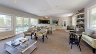 This single-floor community clubhouse offers a thoughtfully designed gathering space that balances comfort and functionality. The main lounge area is generously proportioned, featuring plush seating arrangements, recessed lighting, and large windows that flood the interior with natural light and frame views of lush greenery and an outdoor pool. A fireplace anchors one end of the lounge, complete with a classic white mantel, stone surround, and a wall-mounted television above, creating an inviting focal point. The open layout flows seamlessly into a dining area outfitted with tables and chairs, ideal for group gatherings and events. Adjacent to the dining space, the fully equipped kitchen showcases dark cabinetry, granite countertops, a mosaic tile backsplash, and black appliances, providing everything needed to support catered or self-serve functions. A dedicated game and activity area features casual seating, a round game table, and additional lounge chairs bathed in natural light from oversized windows overlooking the wooded surroundings. Throughout the space, neutral tones, crown molding, and carefully chosen artwork contribute to a polished yet welcoming ambiance, making this clubhouse an ideal venue for both relaxed socializing and organized community activities.