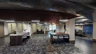 Located at 1301 N Speer Blvd in Denver, Colorado, this contemporary commercial office suite offers a thoughtfully designed, single-floor workspace that balances open collaboration with private meeting areas. The layout is defined by a central open workspace featuring patterned carpet tile flooring, a standout slatted wood ceiling with pendant lighting, and a communal high-top table ideal for team gatherings or informal meetings. Exposed industrial-style ductwork adds an architectural contrast to the refined wood and neutral finishes found throughout. Glass-enclosed conference rooms line the perimeter, providing visual transparency while maintaining acoustic privacy. These meeting rooms are equipped with seating for small to mid-sized groups and are accented with framed artwork and soft interior lighting. Wide, dark-tiled corridors connect the various areas of the suite, creating clear circulation paths between the open workspace and enclosed rooms. Built-in shelving niches and glass-paneled entry doors contribute to the polished, professional aesthetic. A dedicated print and support station is conveniently positioned within the open area. The overall ambiance is modern and professional, combining warm wood tones, cool gray flooring, blue accent walls, and clean architectural lines to create a functional yet visually engaging office environment.