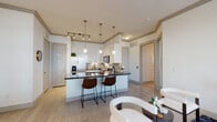 This single-floor residence is thoughtfully designed with a contemporary aesthetic, combining functionality and style across its well-defined living spaces. The open-concept kitchen and living area serves as the heart of the home, where a sleek kitchen features crisp white cabinetry, dark granite countertops, stainless steel appliances including a French door refrigerator, electric range, and over-the-range microwave. A subway tile backsplash adds a clean, modern touch, while a peninsula with seating for two provides a casual dining option beneath elegant brass pendant lights. Light wood-toned flooring extends throughout, unifying the space.

The living area offers generous room for comfortable seating arrangements, with large windows dressed in white curtains and blinds allowing natural light to fill the space. Crown molding with subtle accent detailing borders the ceilings, adding a refined architectural touch.

The primary bedroom includes direct access to a walk-in closet and a private en-suite bathroom featuring a soaking tub, shower, white cabinetry, and dark granite vanity. A secondary bedroom, compact yet functional, features a window and light-toned walls, accommodating flexible uses. The shared bathroom mirrors the same refined finishes found throughout the residence, maintaining a cohesive, modern design.
