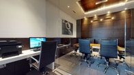 This contemporary office space is thoughtfully designed to balance focused individual work with collaborative functionality. The layout is divided into two distinct zones, separated by a full-height glass partition that maintains an open, airy feel while providing acoustic and visual separation between areas.

The workstation area features a clean white desk equipped with a desktop computer, paired with a stylish black leather executive chair with chrome accents. Dark wood cabinetry lines the walls, providing ample storage while adding warmth and depth to the space.

The adjacent conference area centers around a wood-toned meeting table surrounded by matching black leather executive chairs, accommodating small team discussions or client meetings. A striking black-and-white photographic artwork adorns the white upper wall, adding a refined artistic touch.

The ceiling design is a standout architectural feature, incorporating a coffered tray detail finished in rich dark wood with integrated warm LED strip lighting, creating a dramatic layered effect. Recessed spotlights throughout provide even, professional illumination. Dark wood wall paneling in the conference zone further reinforces the upscale, executive aesthetic. The overall ambiance is polished, professional, and modern — well-suited for a high-end corporate environment.