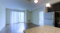 This single-floor space showcases a clean, modern design with an open-concept layout that seamlessly connects the kitchen and living areas. The living space is anchored by rich hardwood flooring that runs throughout, reflecting natural light and adding warmth to the interior. Large sliding glass doors fitted with vertical blinds allow generous amounts of natural light to flood the space, while also providing access to an outdoor area. The kitchen is highlighted by elegant granite countertops in a warm beige and gray tone, paired with contemporary flush-mount and pendant lighting fixtures on the ceiling. White walls and bright trim give the space a crisp, airy feel, while HVAC vents integrated into both the ceiling and the lower wall ensure year-round comfort. A hallway off the main living area leads to additional rooms, suggesting a well-organized floor plan. The overall ambiance is bright, open, and contemporary, making it an inviting and functional living environment suited for a variety of lifestyles.