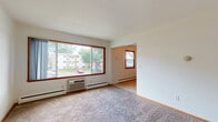 This single-floor unit offers a practical and straightforward layout with clearly defined living spaces. The living room is generously sized, featuring plush carpeting, a large picture window framed in warm wood trim, and a wall-mounted air conditioning unit paired with baseboard heating, ensuring year-round comfort. Natural light fills the space, offering views of the surrounding greenery and neighboring buildings. A secondary carpeted room, currently unfurnished, provides flexible use as a bedroom or additional living area, complete with a ceiling fan and dual windows that invite further natural light and outdoor views.

The kitchen is efficiently laid out with white cabinetry extending to the ceiling for ample storage, a stainless steel sink, an electric range, and a dishwasher. Light tile flooring and a ceiling exhaust fan complete the functional space. Adjacent to the kitchen, a wood-trimmed door leads to additional areas of the unit.

The bathroom features classic mosaic tile flooring in warm earth tones, a white vanity with a mirrored medicine cabinet, a toilet, and a full tub-and-shower combination with white tile surround. Warm wood door frames throughout the unit add a cohesive, understated character to the overall interior.