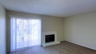 This single-floor residence offers a straightforward and functional layout designed for comfortable everyday living. The living area serves as the focal point of the space, featuring wall-to-wall carpeting that adds warmth underfoot and a clean, neutral color palette throughout. A standout feature of the room is the fireplace, framed by a simple white surround and raised hearth, providing both a visual anchor and a cozy gathering spot. Natural light fills the space generously through a large sliding glass door fitted with vertical blinds, which also offers a view of the outdoors and direct access to an exterior area. The walls are finished in a soft, neutral tone that complements the overall understated aesthetic, while the textured popcorn ceiling is consistent with the classic residential construction style. The open feel of the living area, combined with the abundance of natural light and the warmth of the carpeted flooring, creates an inviting atmosphere. The space is clean and move-in ready, providing a solid canvas for personalization and interior design.