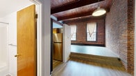 Located at 1 Union Pl in Hartford, Connecticut, this single-floor loft-style unit showcases a compelling blend of industrial heritage and contemporary updates. The space is defined by its striking exposed brick walls, which span the full height of the rooms and lend a warm, textured aesthetic throughout. Rich, dark wooden beams cross the ceilings, complementing the light hardwood flooring found across the main living areas.

The open main living area benefits from generous natural light, courtesy of multiple large windows set within the brick walls, dressed with sheer curtains that soften the industrial character of the space. The layout includes a distinct kitchen area outfitted with warm wood cabinetry, stainless steel appliances including a refrigerator and range, and pendant lighting overhead. A bathroom features wood-toned cabinetry, a single vanity with a large mirror, and clean white finishes.

The bedroom area is a spacious, open room with light flooring and white walls, contrasted by the exposed beams above. Throughout the unit, the interplay of raw brick, timber, and updated finishes creates a cohesive urban loft ambiance well-suited to city living in downtown Hartford.