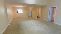 This two-story home offers a generous, open layout with neutral beige walls and soft wall-to-wall carpeting that creates a warm, understated atmosphere throughout. The main living area is notably expansive, providing ample room for flexible furniture arrangements. An elegant chandelier serves as a focal point, adding a touch of refinement to the open space. Natural light filters in through a window, brightening the room with a soft, welcoming glow. The elevated ceiling height enhances the sense of openness, contributing to an airy feel. Adjacent to the main living area, a doorway leads to an en-suite bathroom, which features a toilet and what appears to be a walk-in closet, offering practical convenience. A secondary doorway provides access to an additional room, suggesting a well-connected floor plan designed for ease of movement. The clean, neutral color palette throughout the space makes it highly adaptable to a variety of interior design styles. Overall, the home presents a straightforward, functional layout with comfortable finishes suited for everyday living.