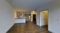 This single-floor unit offers a clean, modern aesthetic with an open-concept layout that seamlessly connects the kitchen and living areas. The kitchen is a standout feature, showcasing light wood cabinetry, granite countertops, and a full suite of stainless steel appliances including a refrigerator, stove, and microwave. A peninsula island provides additional workspace and casual seating, while pendant lighting and a track light fixture above add focused illumination to the area. Rich, dark hardwood-style flooring runs throughout the main living space, adding warmth and a sense of continuity to the open floor plan. The layout flows naturally from the kitchen toward the entry foyer, which is accessible through a solid dark wood front door equipped with a keypad entry system. A side corridor near the entry hints at additional rooms or storage beyond the main living area. The neutral wall tones and recessed ceiling details contribute to a bright, inviting atmosphere. Overall, the space presents a well-designed, functional interior that balances contemporary style with everyday practicality, making it an appealing and comfortable living environment.