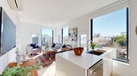 This contemporary single-floor residence showcases a bright, modern aesthetic defined by clean lines, white walls, and light hardwood flooring throughout. The open-plan layout seamlessly connects the kitchen and living area, creating a spacious and airy atmosphere. Floor-to-ceiling windows on multiple sides flood the interior with natural light and frame sweeping urban rooftop views, giving the space an elevated, connected-to-the-city feel.

The kitchen is a standout feature, offering sleek white cabinetry, a bold cobalt blue penny-tile backsplash, stainless steel appliances, and a generous white quartz island with seating for multiple guests — ideal for both everyday living and entertaining. A large window above the sink further enhances the sense of openness.

The living area is generously proportioned, anchored by oversized windows that wrap the corner and invite abundant sunlight throughout the day. The bedroom is a calm, well-appointed retreat featuring a wall-mounted mini-split system for climate control and a recessed ceiling light. A compact, well-finished bathroom with warm wood-toned flooring and clean white finishes completes the layout. Every corner of this residence reflects thoughtful modern design with an emphasis on light, views, and livability.
