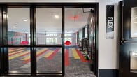 This contemporary fitness facility in Birmingham, Alabama, is designed to energize and inspire with its bold aesthetic and well-equipped workout zones across a single thoughtfully laid-out floor. The space makes an immediate visual impression through its striking multi-colored carpet tiles — featuring bold stripes of red, yellow, and grey — that visually define each area and contribute to the facility's dynamic, high-energy atmosphere.

The main workout area is expansive and open, flooded with natural light from a series of large windows that offer views of surrounding greenery. A full-wall mirror and ballet barre create a dedicated studio zone, complemented by a textured gold accent wall adorned with abstract line art. Cardio equipment including stationary bikes and elliptical machines lines the perimeter, while a well-stocked rack of kettlebells, medicine balls, and stability balls supports functional and strength training.

A separate open-floor exercise room features a bold mural, suspended training accessories, and a railing-lined open wall that brings in additional natural light. The facility's entry area is finished with a rustic stacked-stone accent wall and a dramatic red statement wall flanking dark double doors, setting a confident, energetic tone from the moment of arrival. Recessed lighting throughout ensures a well-lit environment at all times.