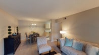 This single-floor, 2-bedroom, 2-bathroom residence offers a well-appointed and functional layout designed for comfortable living. The open-concept living and dining area creates a seamless flow, anchored by warm, neutral tones and plush carpeting that add a cozy ambiance throughout. The living area provides generous space for seating arrangements, complemented by soft, inviting lighting. Adjacent to the living space, the dining area features a dedicated area for a full dining set, accented by an elegant chandelier that adds a refined touch. A large decorative wall clock adds a stylish focal point to the dining space. The kitchen, visible from the dining area, features light wood cabinetry and ample counter space, offering both practicality and a clean, bright aesthetic. A half-wall partition subtly defines the transition between the living and dining zones while maintaining an open feel. The textured ceiling adds architectural character throughout the main living areas. With two bedrooms and two full bathrooms, this residence provides privacy and convenience. The neutral color palette and warm lighting create an inviting atmosphere that is both functional and stylish, making it an ideal space for everyday living.