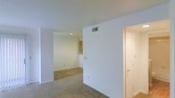 This single-floor residential space offers a clean, functional layout designed for comfortable everyday living. The interior is finished with neutral white walls and smooth ceilings throughout, creating a bright and airy atmosphere. The living area features wall-to-wall carpeting that adds warmth underfoot, while recessed lighting provides consistent illumination across the open floor plan. A wide doorway connects the main living space to an adjacent area, promoting a seamless flow between rooms. Visible from the living space, the kitchen is outfitted with warm-toned wood cabinetry and a raised counter or bar-height divider, offering both workspace and a casual dining option. HVAC vents are thoughtfully integrated into the ceiling and upper walls, ensuring efficient climate control throughout. A doorway leading to an additional room or entryway is visible to the right, hinting at further functional spaces within the unit. The overall ambiance is understated and move-in ready, with a simple, well-maintained interior that provides a neutral canvas for personalization.