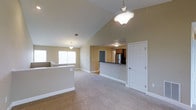 This single-floor residence offers a welcoming open-concept layout designed for both comfort and functionality. The living area immediately impresses with its vaulted ceilings that rise dramatically overhead, creating a generous sense of vertical space and airiness. Warm neutral-toned walls and plush carpeting throughout lend the space a cozy, inviting ambiance. Pendant light fixtures hang gracefully from the high ceiling, providing both practical illumination and decorative character.

Natural light pours in through a wide window at the far end of the living area, brightening the room and enhancing its open feel. A half-wall partition defines the living space while maintaining the flow of the open floor plan. The kitchen is positioned toward the rear and features dark countertops, wood-toned cabinetry, and a stainless steel refrigerator, offering a functional and attractive workspace. A raised breakfast bar serves as a natural transition between the kitchen and the main living area, ideal for casual dining or entertaining.

A paneled white door provides access to an adjacent space, adding to the home's practical layout. Overall, the design balances open, airy living with defined functional zones, making it a versatile and comfortable place to call home.