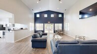 Located at 202 Lakeview Park Rd in Colonial Heights, Virginia, this single-floor space offers a well-designed, open layout ideal for gatherings and community use. The main area immediately impresses with its dramatic vaulted ceilings that rise to a striking peak, creating a sense of volume and grandeur. A bold navy blue accent wall serves as a focal point, complemented by crisp white walls and warm wood-look flooring throughout. A row of clerestory windows near the roofline, paired with large windows and glass French doors along the front wall, floods the interior with abundant natural light and offers views of the surrounding wooded landscape. The main room is thoughtfully arranged with seating and a dining area, while a wall-mounted TV and a built-in shelf add functional detail. French doors open to a secondary area with additional workspace, accessed through mirrored panel doors that enhance the sense of depth. The kitchen is clean and modern, featuring white shaker cabinetry, granite-look countertops, stainless steel appliances including a range, microwave, and refrigerator, and a double-basin sink beneath a window. The overall ambiance is bright, airy, and welcoming, blending contemporary finishes with a comfortable, functional design.